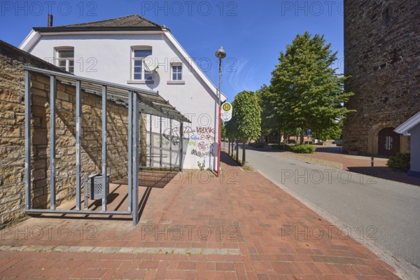 Bus stop church of the lines 670 Rieste and 672 Thiene, bus shelter, building, lantern, church tower, quarry stone wall, graffiti, trees, blue sky, cirrostratus clouds, main street, Alfhausen, district of Osnabrück, Lower Saxony, Germany