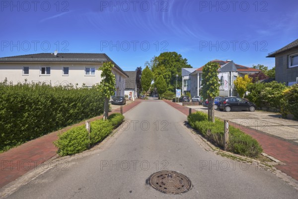 Multi-family houses, general development, residential building, trees, hedge, clinker pavement, asphalt road, manhole cover, road with narrowed carriageway, traffic-calmed area, blue sky, cirrostratus clouds, main road, Alfhausen, district of Osnabrück, Lower Saxony, Germany