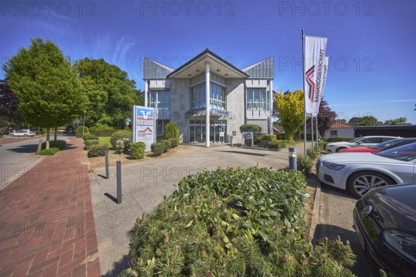 Vereinigte Volksbank eG - Filiale Alfhausen, Baugenossenschaft Landkreis Osnabrück eG, flags on flagpoles, modern architecture, street, car park with vehicles, bollards, flower bed, trees, hedge, blue sky, cirrostratus clouds, Hauptstraße, Alfhausen, Landkreis Osnabrück, Lower Saxony, Germany