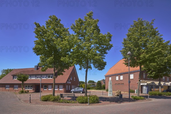 Street, bollards, trees, bench, 1000 year fountain, lantern, clinker brick pavement, concrete paving stone street, shady, pavement, clinker brick building, hedges, blue sky, cloudless, intersection of Hauptstraße with Nonnenpfad and In den Gärten, Alfhausen, Osnabrück district, Lower Saxony, Germany