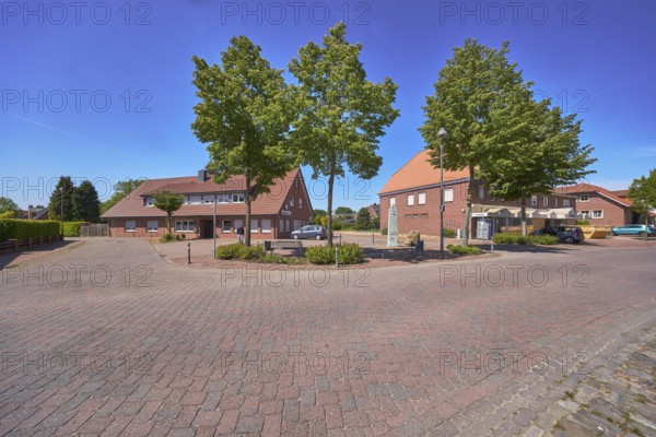 Street, bollards, trees, bench, 1000 year fountain, lantern, clinker brick pavement, concrete paving stone street, shady, pavement, clinker brick building, hedges, blue sky, cloudless, intersection of Hauptstraße with Nonnenpfad and In den Gärten, Alfhausen, Osnabrück district, Lower Saxony, Germany