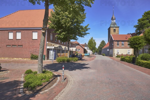 Street, general development, clinker brick building, pointed roof with red roof tiles, church tower of St. Johannis, trees, clinker brick pavement, asphalt road, pavement, blue sky, cirrostratus clouds, intersection of Hauptstraße and Nonnenpfad, Alfhausen, district of Osnabrück, Lower Saxony, Germany