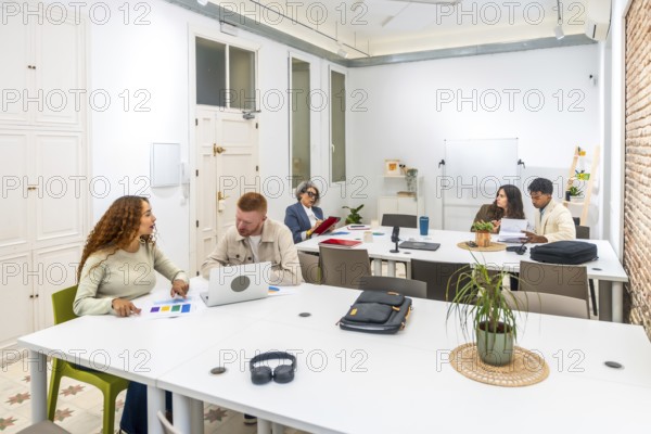 Group of diverse business professionals discussing ideas, analyzing documents, and working on laptops during a collaborative meeting in a bright, modern office space