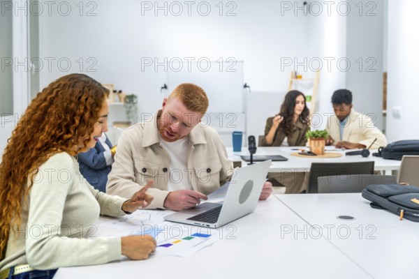 Young diverse professionals collaborating at a meeting table, discussing business strategies, analyzing data charts, and using a laptop in a modern office environment