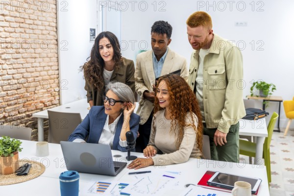 Diverse business team working together in a modern office, grouping around a laptop and microphone, discussing data charts and sharing ideas during a casual meeting
