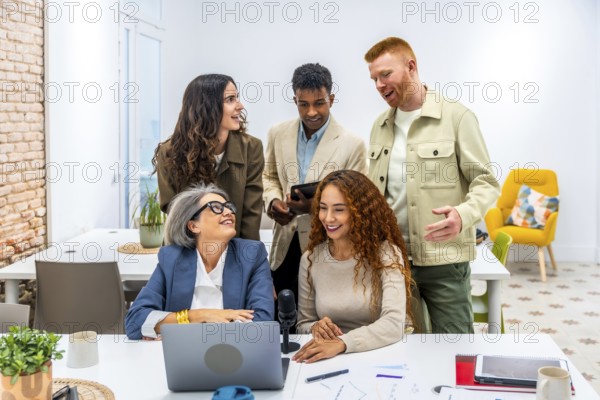 Diverse business team collaborating and brainstorming ideas during a casual work meeting in a modern office, using a laptop, tablet, and microphone for a podcast or online discussion