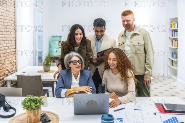 Multigenerational and diverse business team members working together, discussing ideas, and looking at a laptop screen in a bright co working office space, fostering collaboration