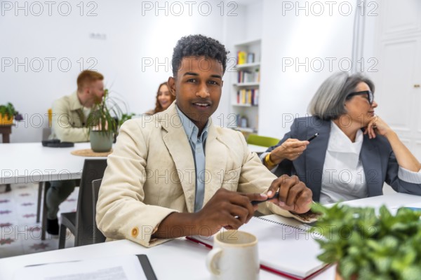Young african american man smiling at the camera during a business meeting, with other diverse colleagues working and discussing in the background, embracing teamwork and inclusion