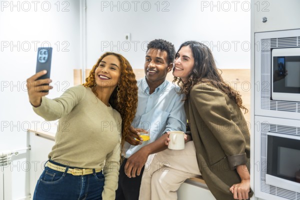 Group of diverse colleagues enjoying a coffee break in the office kitchen, smiling and posing together for a selfie using a smartphone, showing friendship and teamwork