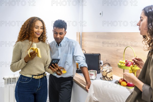 Colleagues from diverse backgrounds taking an office break in a contemporary kitchen, snacking on fresh fruit and engaging with a smartphone while socializing