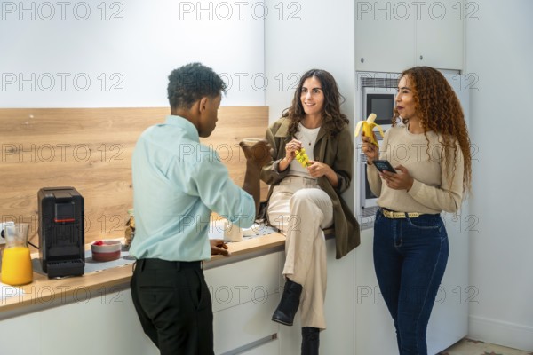 Diverse colleagues chatting and smiling during a casual office kitchen break, two women holding fruit and a smartphone while a male coworker talks, relaxed teamwork vibe
