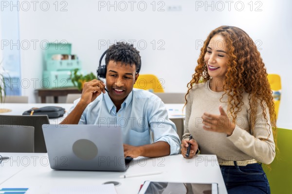 Diverse colleagues at an office desk collaborate as a man with a headset uses a laptop while a woman supports and communicates, smiling and providing customer service and technical help