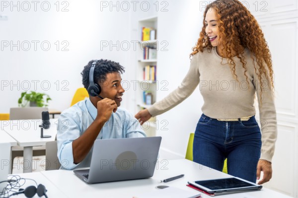 Multiracial coworkers smiling and interacting in a modern office, with a man wearing a headset using his laptop while a woman stands nearby, discussing work