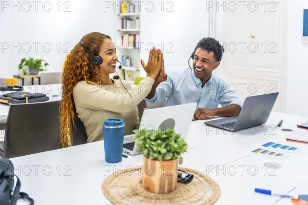 Two cheerful multiethnic customer service agents wearing headsets giving a high five, celebrating a successful outcome while working on laptops in a modern office