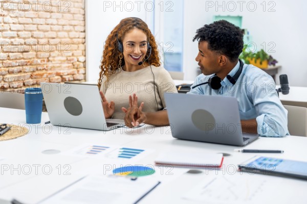 Diverse male and female coworkers with headsets collaborate at laptops on a bright modern office desk, providing customer support, training and online assistance as a team