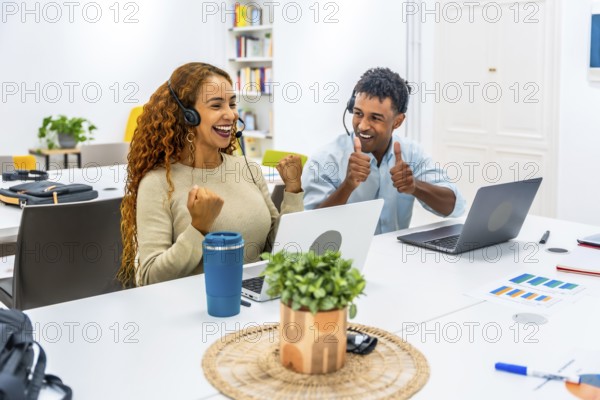 Two diverse call center agents wearing headsets and working on laptops, celebrating their achievement with joy, showing thumbs up and a triumphant fist in a bright modern office