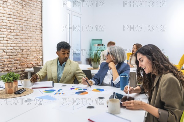 Multiracial business professionals discussing colorful charts and data analytics during a collaborative meeting, working together on a project in a bright, contemporary office environment
