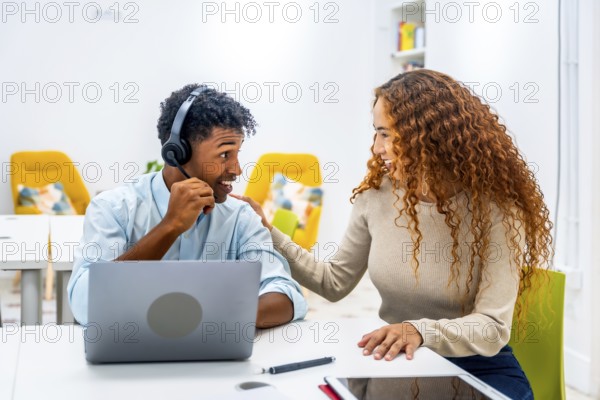 Two diverse colleagues in a modern office setup, a man wearing a headset using a laptop while a woman offers support and guidance, highlighting teamwork and effective communication