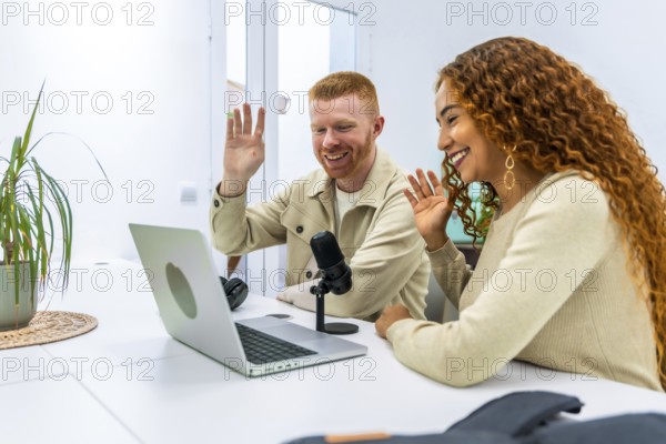 Diverse young adult man and woman smiling and waving while actively recording a podcast or participating in a video call event from home, using a laptop, microphone, and headphones
