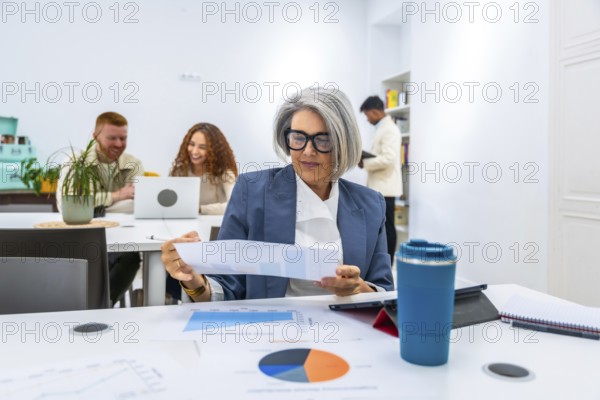 Mature female manager reviewing a financial report with charts and graphs on a table, collaborating with diverse young coworkers in a modern office workplace