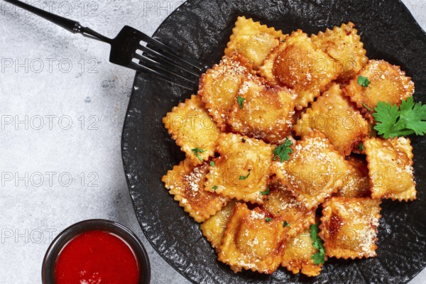 Fried ravioli with parmesan cheese, served with red sauce and garnished with herbs, on a black plate, no people