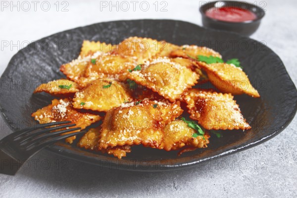 Fried ravioli with parmesan cheese, served with red sauce and garnished with herbs, on a black plate, no people
