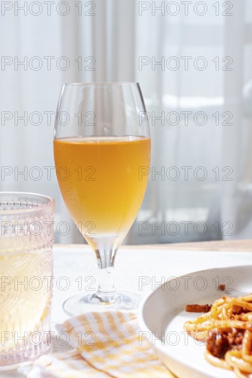 A glass filled with a light amber drink sits beside a plate of pasta. Bright sunlight streams through sheer curtains, creating a warm and inviting atmosphere