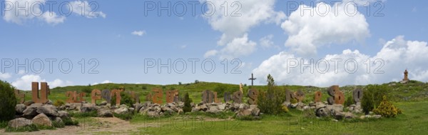 Wide landscape with rocks and a cross, surrounded by green meadow and blue sky, monument of the Armenian alphabet, Aparan, Aragazotn province, Armenia