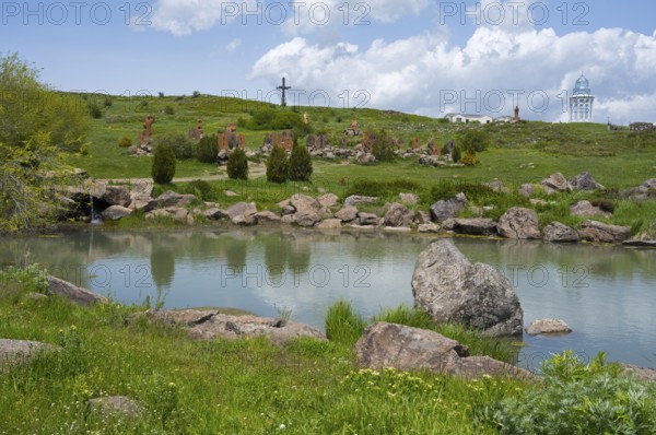 Peaceful lake surrounded by rocks and green landscape with a cross on a hill, monument of the Armenian alphabet, Aparan, Aragazotn province, Armenia