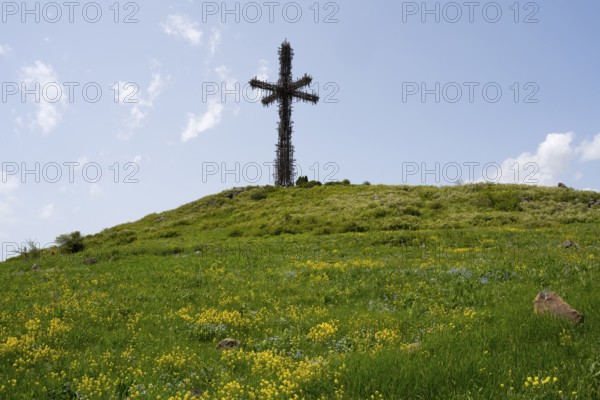 Metal cross on a green hill under a cloudy blue sky, surrounded by grass and yellow flowers, Monument of the Holy Cross, consisting of many small metal crosses, Aparan, Aragazotn Province, Armenia
