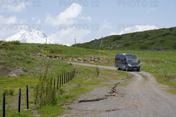 A car drives on a rural road in front of a snow-covered mountain under a blue sky, camper drives past the Garden of Eternity, in the background the Aragaz, Aparan, Aragazotn Province, Armenia