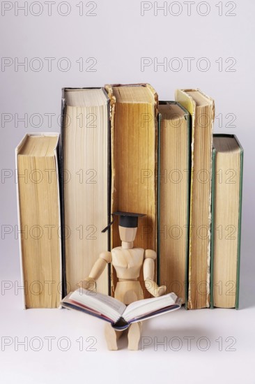 A wooden man in a student cap sits against a background of a stack of textbooks and reads a book, space for text