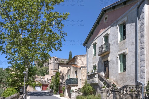 The sunlit village street of Moureze is lined with traditional stone houses with reddish-brown garage doors, while the medieval bell tower and the craggy cliffs of the Cirque de Moureze loom in the background: Moureze, Grand Site Salagou-Cirque de Moureze, Herault, Occitania, France