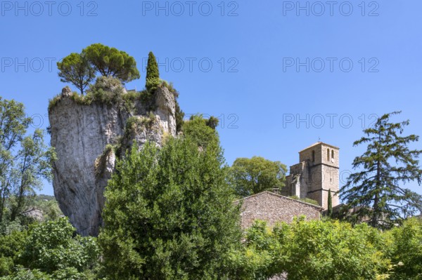 The Romanesque church of Sainte-Marie with its massive bell tower stands embedded in the green of the foreground, overlooked by the spectacular, eroded dolomite rock Roc Castel in the heart of the rocky circus of Moureze, Grand Site Lac du Salagou, Cirque de Moureze, Herault, Occitania, France