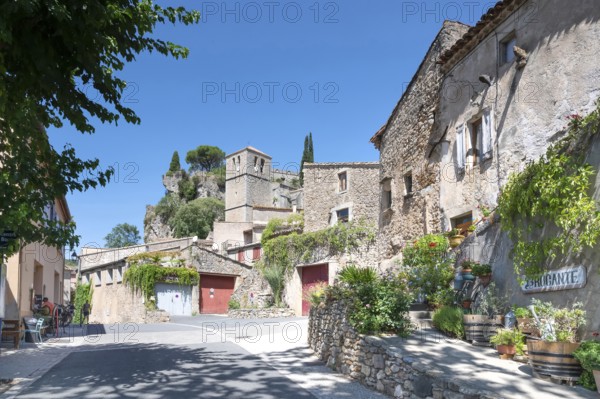 The sunlit village street of Moureze is lined with traditional stone houses with reddish-brown garage doors, while the medieval bell tower and the craggy cliffs of the Cirque de Moureze loom in the background: Moureze, Grand Site Salagou-Cirque de Moureze, Herault, Occitania, France
