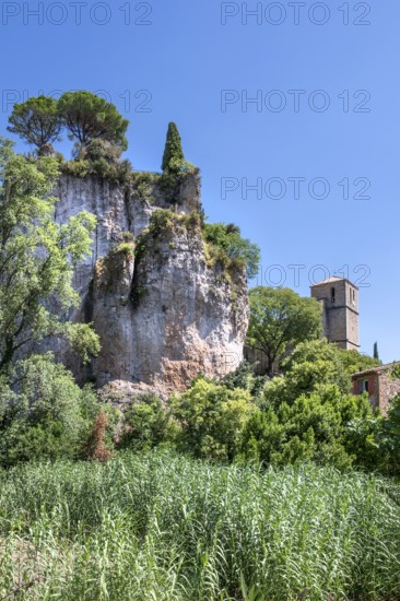 The Romanesque church of Sainte-Marie with its massive bell tower stands embedded in the green of the foreground, overlooked by the spectacular, eroded dolomite rock Roc Castel in the heart of the rocky circus of Moureze, Grand Site Lac du Salagou, Cirque de Moureze, Herault, Occitania, France