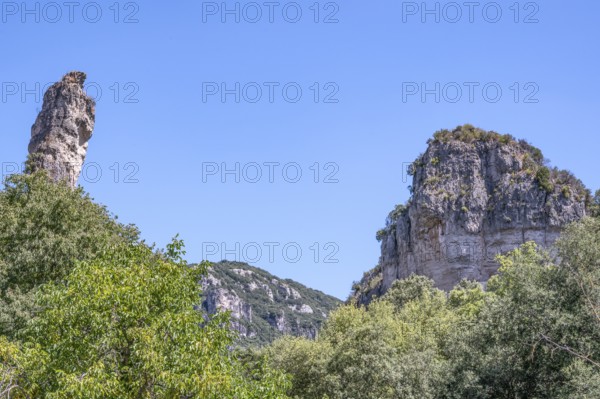 Two prominent dolomite cliffs formed by erosion - a towering peak on the left and a massive rock face on the right - above lush green vegetation under a blue sky, typical of the Cirque de Moureze: Moureze, Herault, Occitania, France