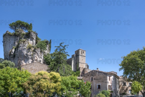 The Romanesque church of Sainte-Marie with its massive bell tower stands embedded in the green of the foreground, overlooked by the spectacular, eroded dolomite rock Roc Castel in the heart of the rocky circus of Moureze, Grand Site Lac du Salagou, Cirque de Moureze, Herault, Occitania, France