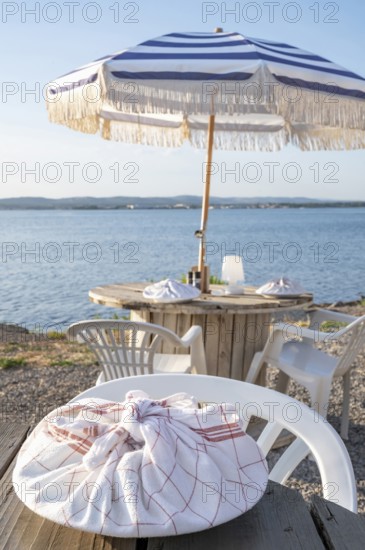 A plate wrapped in a red and white chequered tea towel lies on a rustic wooden table in the foreground at the beach restaurant Guinguette a la Bonne Franquette, with a blurred view of the glistening waters of the Etang de Thau, a round dining table with parasol and the busy coastline in the background, Sete, Herault, Occitania, France