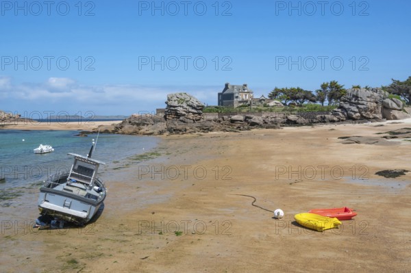 On the harbour beach of Tregastel on the Cote de Granit Rose, small boats lie on the light sand at low tide against the backdrop of the iconic pink granite rocks and Breton houses, Tregastel, Cotes-d'Armor department, Brittany region, France