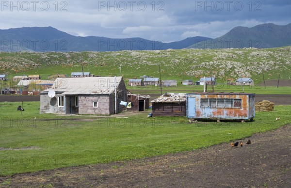 Rustic barns and buildings on green pasture against a mountainous backdrop with clouds in the sky, Shenkani village, Aragazotn province, Armenia