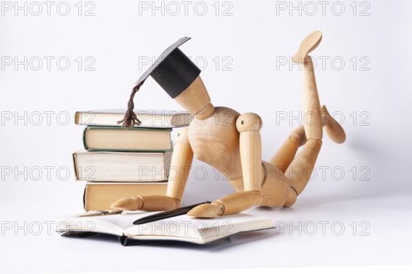 A wooden man in a student hat lies on the floor and reads textbooks
