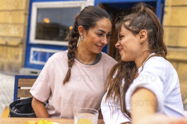Two women are sitting close together at a table outside a cafe, smiling and enjoying each other's company