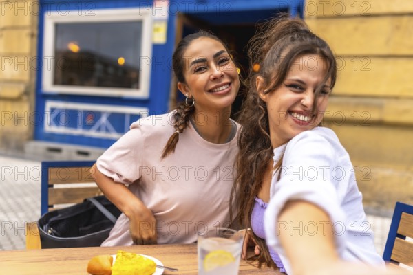 Two friends are taking a selfie while enjoying breakfast at an outdoor cafe, capturing a moment of joy and togetherness during their travel adventure