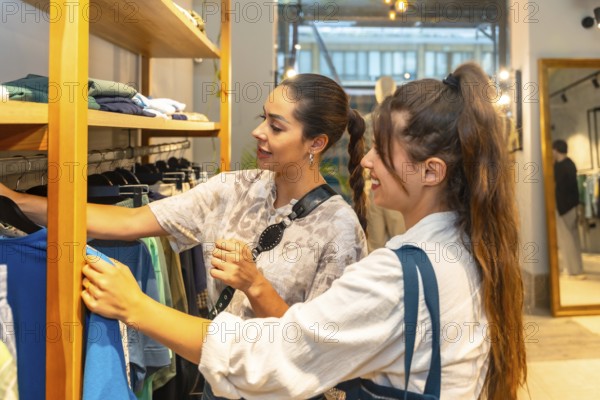 Two young women looking and choosing clothes in a fashion store, the shop assistant is helping a customer