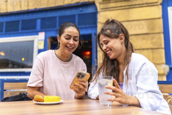 Two female friends are enjoying a moment together at a bar, looking at something funny on a smartphone and laughing