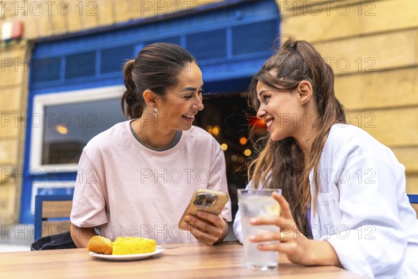 Two women are enjoying a meal and drinks at an outdoor cafe, sharing a moment of connection as they browse a smartphone together