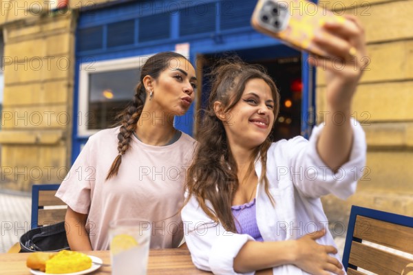 Two young women are taking a selfie with a smartphone while sitting at an outdoor table, enjoying breakfast