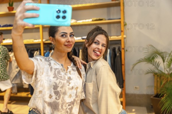 Two cheerful female customers taking a selfie with a smartphone, having fun and making funny faces inside a modern clothing store