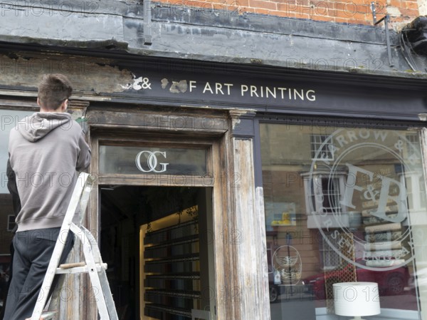Decorator preparing wooden surface of shop front for painting, Devizes, Wiltshire, England, UK